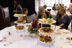 Two towers of food for a high tea and a few people in the back ground sat at round tables